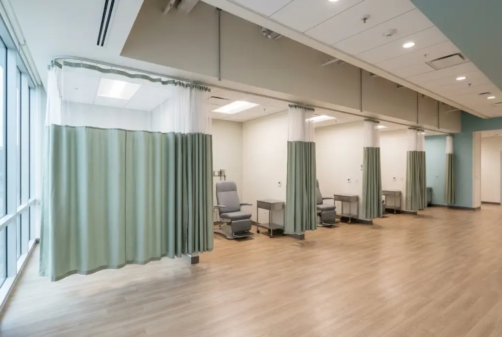 A continuous run of ceiling curtain track along an outpatient treatment floor, dividing six stations behind sage-green mesh-top curtains.